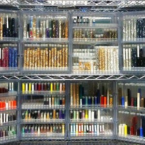 May include: A metal shelving unit filled with clear glass tubes containing various colors of small beads. The beads are organized by color and are arranged in rows on the shelves.