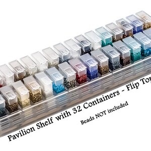 May include: A clear plastic storage organizer featuring a pavilion shelf with 32 individual containers, each equipped with a flip top lid. The text on the base reads Pavilion Shelf with 32 Containers ~ Flip Tops 1-1/2" and Beads NOT included.