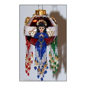 May include: A red glass Christmas ornament with a beaded angel design. The angel is wearing a blue dress and white wings. The ornament is decorated with gold and silver beads and has a beaded fringe.