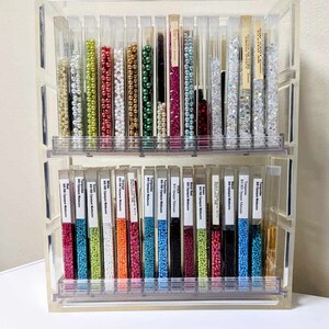 May include: A clear acrylic two-tiered storage organizer filled with vertical tubes of colorful seed beads. Each tube contains a different color, including red, green, blue, white, and metallic tones, with white labels identifying the bead types.