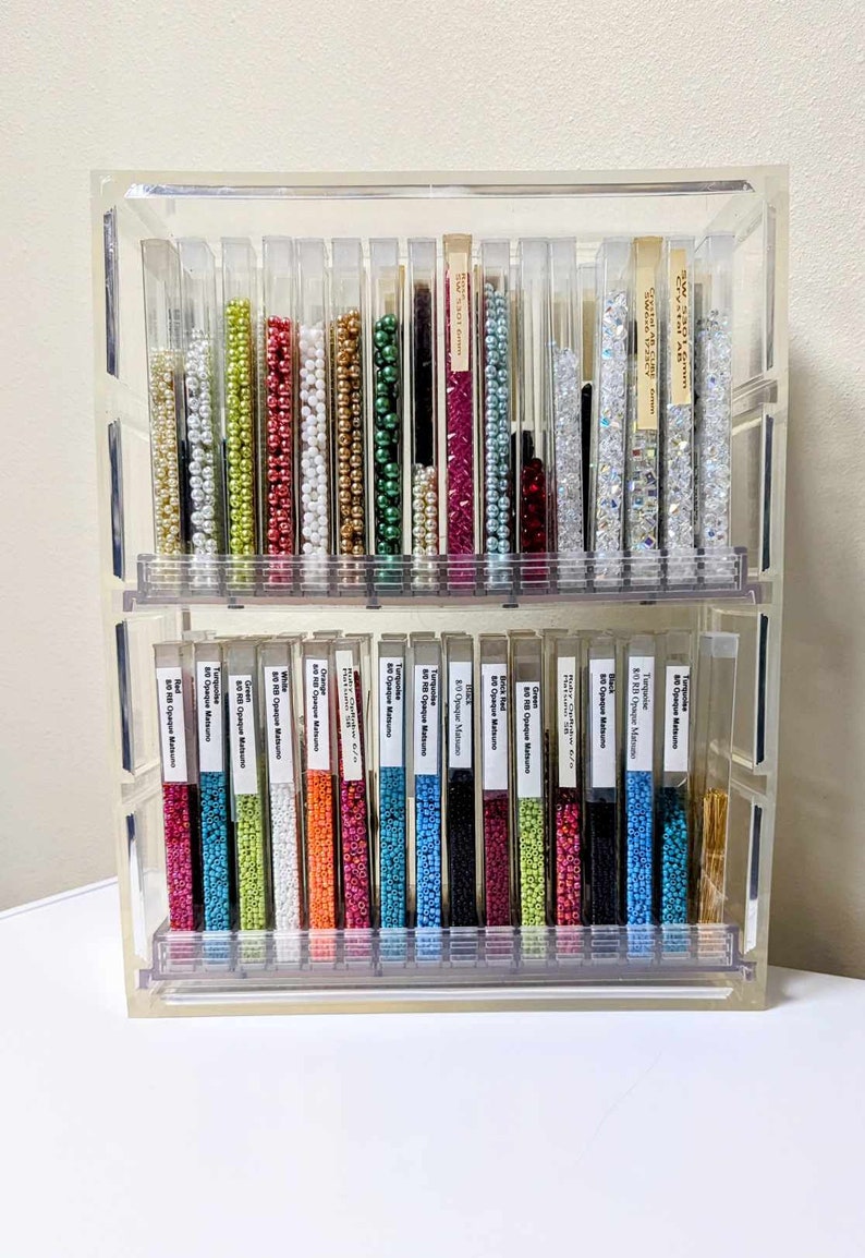May include: A clear acrylic two-tiered storage organizer filled with vertical tubes of colorful seed beads. Each tube contains a different color, including red, green, blue, white, and metallic tones, with white labels identifying the bead types.