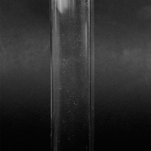 May include: A tall, clear, rectangular container with a white lid and a small, white, folded element extending from the top. The container is transparent, allowing a view of the dark background.