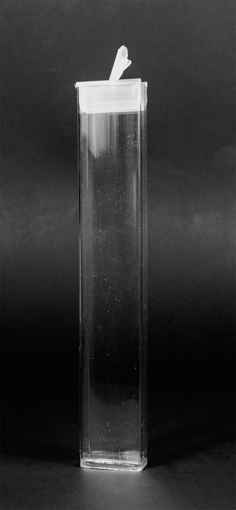 May include: A tall, clear, rectangular container with a white lid and a small, white, folded element extending from the top. The container is transparent, allowing a view of the dark background.