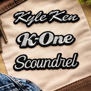 May include: Three embroidered patches with black borders and white lettering. The top patch reads "Kyle Ken", the middle patch says "K-One", and the bottom patch says "Scoundrel". The patches are on a tan fabric.