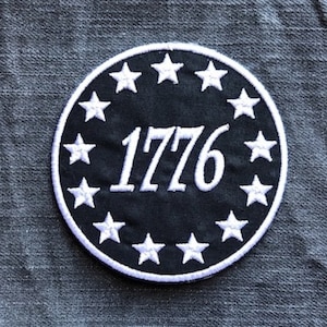 May include: A black and white embroidered patch with a circle of white stars surrounding the number 1776.