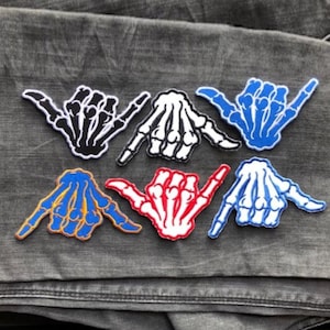 May include: Six embroidered iron-on patches featuring a skeleton hand in a 'hang loose' gesture. The patches are in various colours, including black, white, red, blue, and orange.
