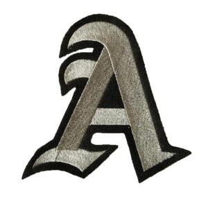 Puede incluir: Un parche de hierro bordado en plata y negro en forma de la letra A. La letra está en una fuente de estilo gótico.