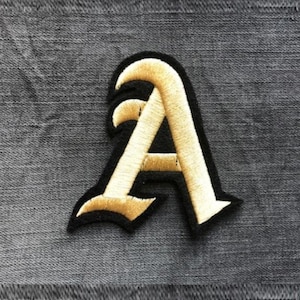 Könnte beinhalten: Ein Bügelbild mit einem goldenen gestickten Buchstaben A im gotischen Schriftstil.