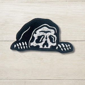 May include: A black embroidered patch featuring a white skull design. The skull is partially covered by a black hood. The patch has a unique shape and is likely designed to be sewn onto clothing or accessories.