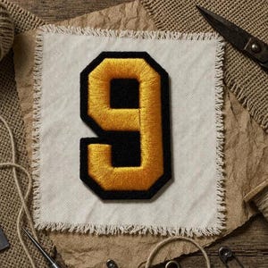 May include: A gold and black embroidered number 9 patch on a white fabric square with frayed edges. The patch is surrounded by sewing tools and materials, including scissors, thread, and pins, on a wooden surface.