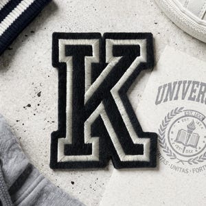 Pode incluir: Um patch com a letra "K" em preto e branco com borda branca, ao lado de um moletom cinza com capuz, uma manga listrada em azul marinho e branco, um tênis branco e um emblema universitário em papel creme.