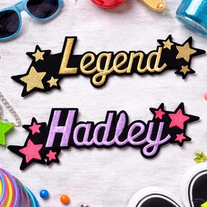 Könnte beinhalten: Zwei gestickte Aufnäher mit den Wörtern "Legend" und "Hadley" in goldener bzw. violetter Schrift auf schwarzen sternförmigen Hintergründen. Die Aufnäher sind von Accessoires wie Sonnenbrillen, Schmuck und Schuhen umgeben.