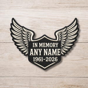 Könnte beinhalten: Schwarzer, gestickter Aufnäher mit weißem Flügeldesign und dem Text "IN MEMORY ANY NAME 1961-2026". Der Aufnäher hat die Form eines Schildes mit schwarzem Rand und ist zum Aufnähen auf Kleidung oder andere Stoffartikel bestimmt.