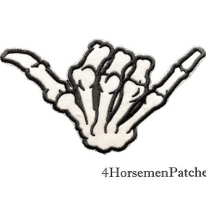 May include: Black and white embroidered iron-on patch of a skeleton hand giving the shaka sign. The patch is for clothing, bags, or other fabric items.