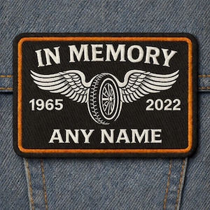 Könnte beinhalten: Schwarzer rechteckiger Aufnäher mit orangefarbenem Rand, mit dem Text „IN MEMORY“ über einem geflügelten Reifendesign. Unter dem Reifen stehen die Jahreszahlen „1965“ und „2022“ sowie der Text „ANY NAME“ unten. Der Aufnäher befindet sich auf einem Jeanshintergrund.