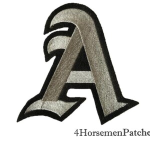 Puede incluir: Un parche de hierro bordado en plata y negro de la letra A en una fuente g&oacute;tica.