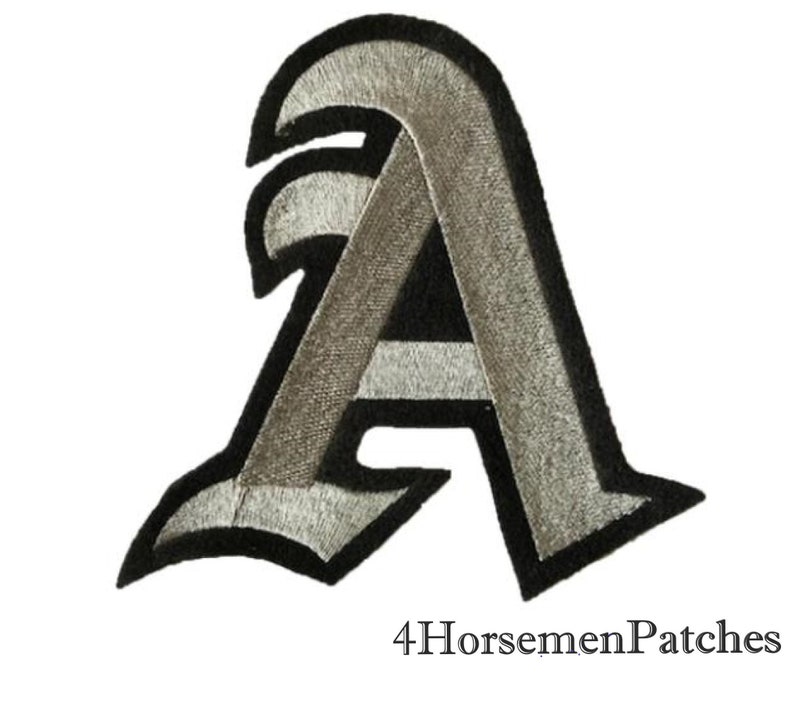 Puede incluir: Un parche de hierro bordado en plata y negro de la letra A en una fuente g&oacute;tica.