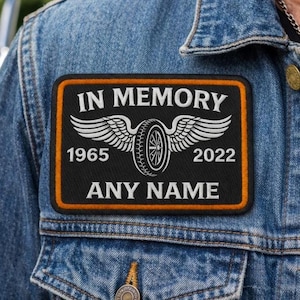 Könnte beinhalten: Ein schwarzer rechteckiger Aufnäher mit orangefarbenem Rand, mit den Worten "IN MEMORY" über einem geflügelten Reifendesign. Die Jahre 1965 und 2022 flankieren den Reifen, darunter Platz für einen Namen. Der Aufnäher ist auf eine Jeansjacke genäht.