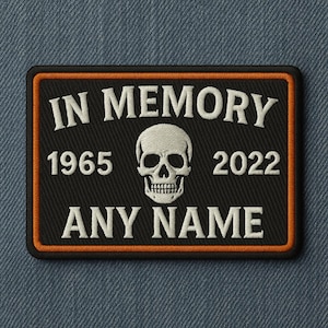 以下が含まれることがあります： オレンジ色の縁取りが付いた黒色の長方形パッチ。「IN MEMORY」の文字が入り、その下に「1965」と「2022」の数字、その間に骸骨のイラストが描かれています。下部には「ANY NAME」の文字。