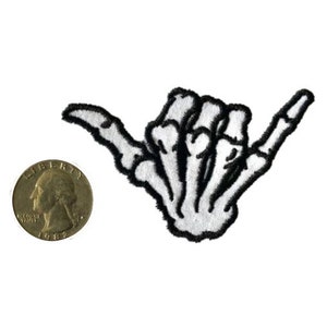 Puede incluir: Parche bordado en blanco y negro de una mano de esqueleto haciendo el signo shaka.