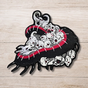 May include: A black and white embroidered patch featuring a cluster of skulls and horned creatures contained within a curved, black, mouth-like shape outlined in red stitching. The design includes multiple small skulls with red accents.