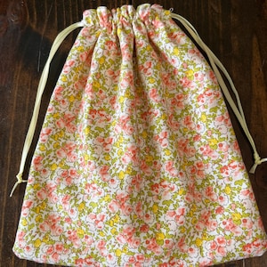 May include: A floral drawstring bag with a pattern of small pink and yellow flowers on a white background. The bag has a gathered top and is tied with yellow ribbon. The bag is made of fabric.