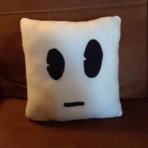 May include: White plush pillow with a black embroidered face. The face has two large oval eyes and a straight line for a mouth.