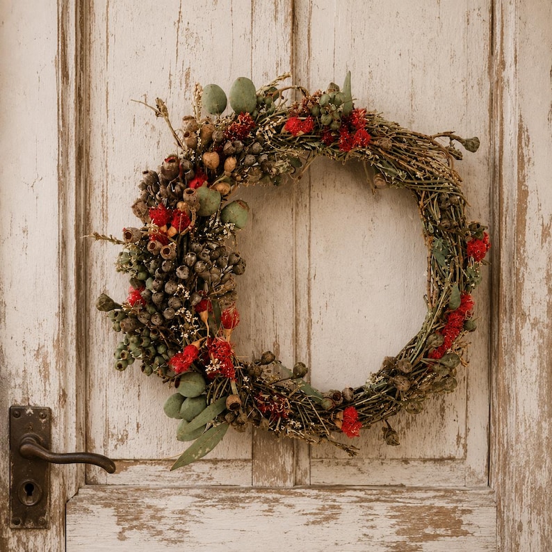 Handmade Australian native dried flower wreath featuring eucalyptus gum nuts, bottlebrush, preserved foliage and rustic twigs, displayed on a weathered white timber door with vintage hardware. An everlasting wreath perfect for front door décor, farmhouse styling, Christmas or Australian home décor