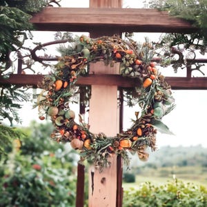 May include: A circular wreath constructed from intertwined brown twigs, decorated with green pine needles, small orange berries, and round green seed pods. The wreath is mounted on a vertical wooden post structure with horizontal crossbeams.
