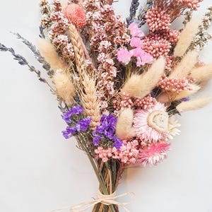 Dried flower bridal bouquet in soft pink, lilac and neutral tones tied with raffia, on a white background.