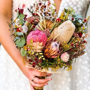 Op de afbeelding: Een boeket droogbloemen met een grote roze protea, een getextureerde beige banksia en diverse kleine rode, gele en groene wilde bloemen. Het arrangement bevat eucalyptusbladeren en is samengebonden met een lichtgekleurd raffialint.