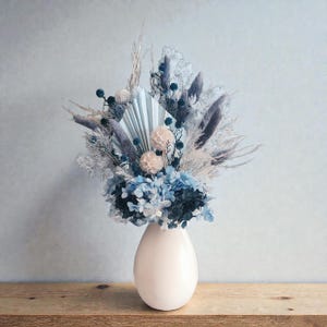 May include: A cream-coloured, egg-shaped vase filled with a dried flower arrangement. The arrangement includes blue hydrangeas, pampas grass, and other dried elements in blue, grey, and beige. The vase is on a wooden surface.