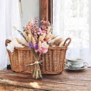 A wicker basket filled with a bouquet of dried flowers, including lavender, wheat, and pink and purple wildflowers. The basket has two handles and is sitting on a wooden surface.