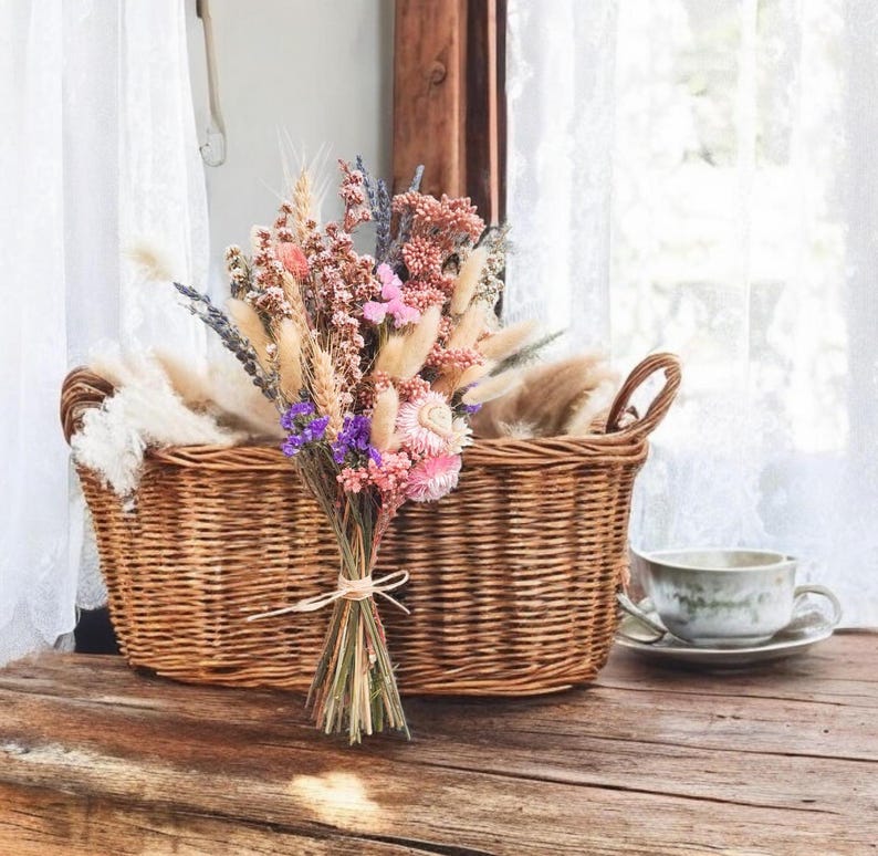 A wicker basket filled with a bouquet of dried flowers, including lavender, wheat, and pink and purple wildflowers. The basket has two handles and is sitting on a wooden surface.