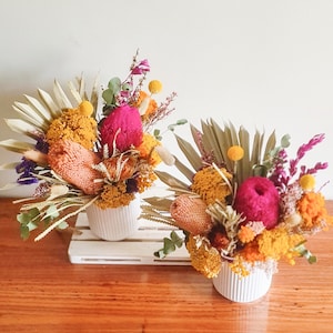 May include: Two white ceramic vases filled with dried flowers. The flowers are a mix of yellow, orange, pink, and purple. The vases are sitting on a white wooden shelf.