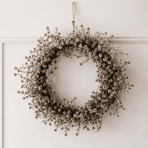 May include: A circular wreath constructed from dried, light brown twigs and numerous small, off-white baby's breath flowers. The wreath is suspended by a thin, light brown twine loop attached to the top, creating a natural, textured appearance.
