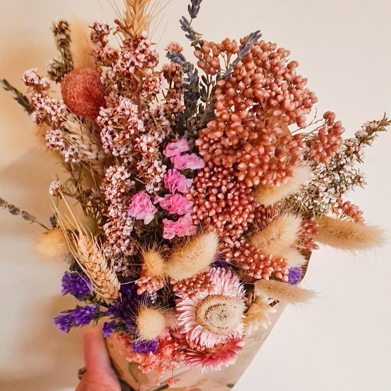 A bouquet of dried flowers in shades of pink, purple, and neutral tones wrapped in brown paper. The bouquet includes dried lavender, wheat, bunny tail grass, everlasting daisies and other dried flowers.