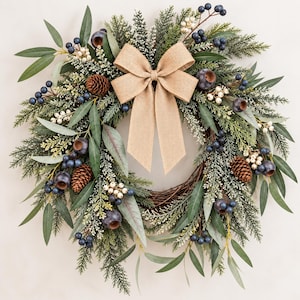 May include: A circular wreath featuring green eucalyptus leaves, evergreen pine branches, and brown pinecones. The arrangement is accented with clusters of dark blue berries and small white berries, centered with a large, tan burlap bow.