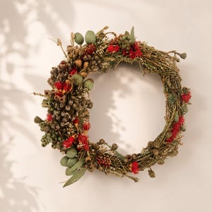 May include: A circular wreath constructed from intertwined brown twigs, decorated with clusters of small green eucalyptus pods, dried brown seed heads, and vibrant red floral accents distributed unevenly around the frame of the wreath.