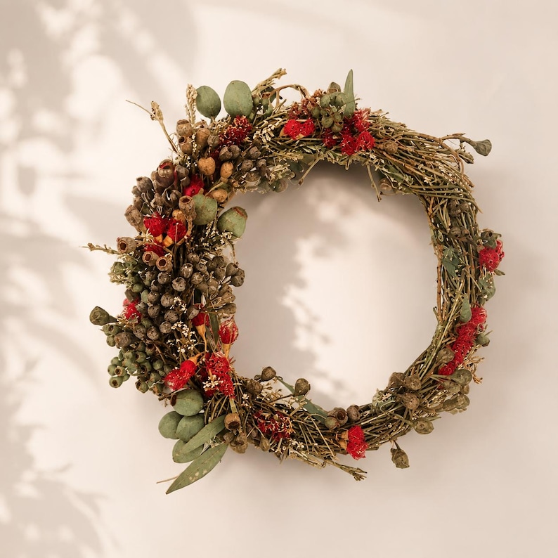 May include: A circular wreath constructed from intertwined brown twigs, decorated with clusters of small green eucalyptus pods, dried brown seed heads, and vibrant red floral accents distributed unevenly around the frame of the wreath.