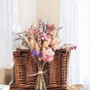 Dried flower bridal bouquet in soft pink, lilac and neutral tones tied with raffia, styled against a wicker basket.