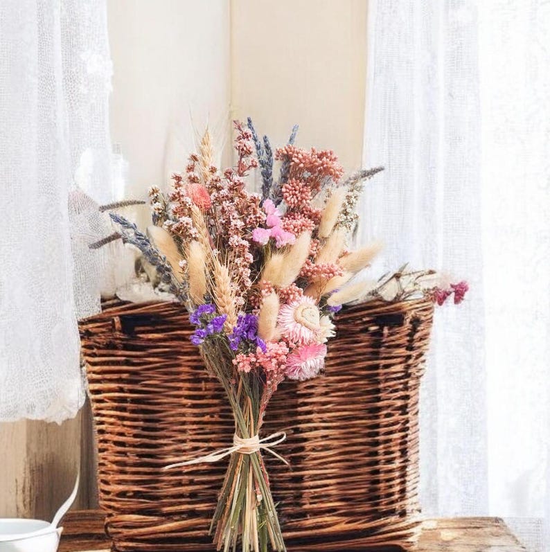 Dried flower bridal bouquet in soft pink, lilac and neutral tones tied with raffia, styled against a wicker basket.