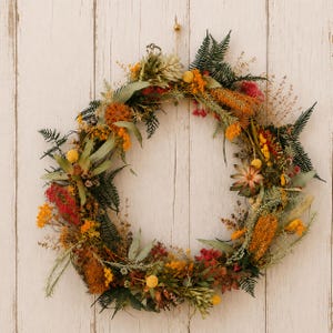 May include: A circular wreath composed of dried flowers and foliage in shades of orange, yellow, red, and green. The arrangement features various textures including fern fronds, eucalyptus leaves, small yellow billy balls, and orange blooms.