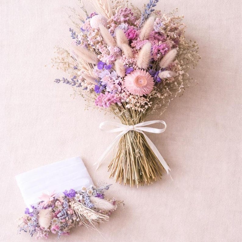 Dried flower bridal bouquet in soft pink, lilac and neutral tones tied with raffia, styled flat-lay on a light linen surface with a matching pocket boutonnière placed beside it.