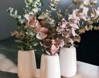 May include: Three white ceramic vases display eucalyptus branches with green, pink, and brown leaves. The vases have a rounded shape and are arranged on a light surface, creating a simple, elegant display.