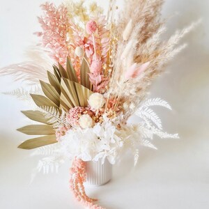 May include: A dried flower arrangement in a white vase. The arrangement features a variety of dried flowers in shades of pink, white, and beige, including pampas grass, dried palm leaves, and fluffy white flowers.