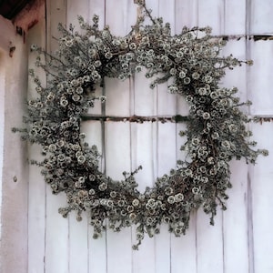 May include: A dried floral wreath in shades of white and gray hangs on a white wooden wall. The wreath is made of small, delicate flowers and branches.