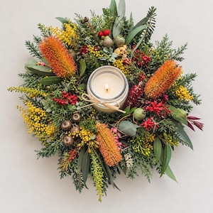 May include: A circular wreath composed of green foliage, yellow mimosa flowers, red berries, and orange banksia blooms surrounding a central glass jar candle with a lit white wick and a decorative tan twine bow tied around the glass rim.