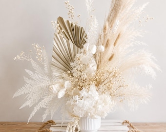 May include: A floral arrangement featuring dried white pampas grass, beige palm spears, white ferns, baby's breath, and hydrangea blossoms. The bouquet is contained in a white ribbed ceramic vase, which sits atop a white wooden tray with rope handles.