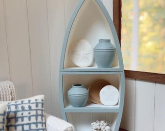 May include: A light blue, boat-shaped wooden shelving unit with three tiers. The shelves contain two light blue vases, two white sand dollar decorations, a small woven bowl, and a white piece of coral, all arranged on white interior panels.